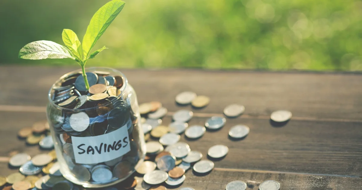 Glass jar filled with coins labeled "savings" with a small green plant growing out of it, symbolizing financial growth.